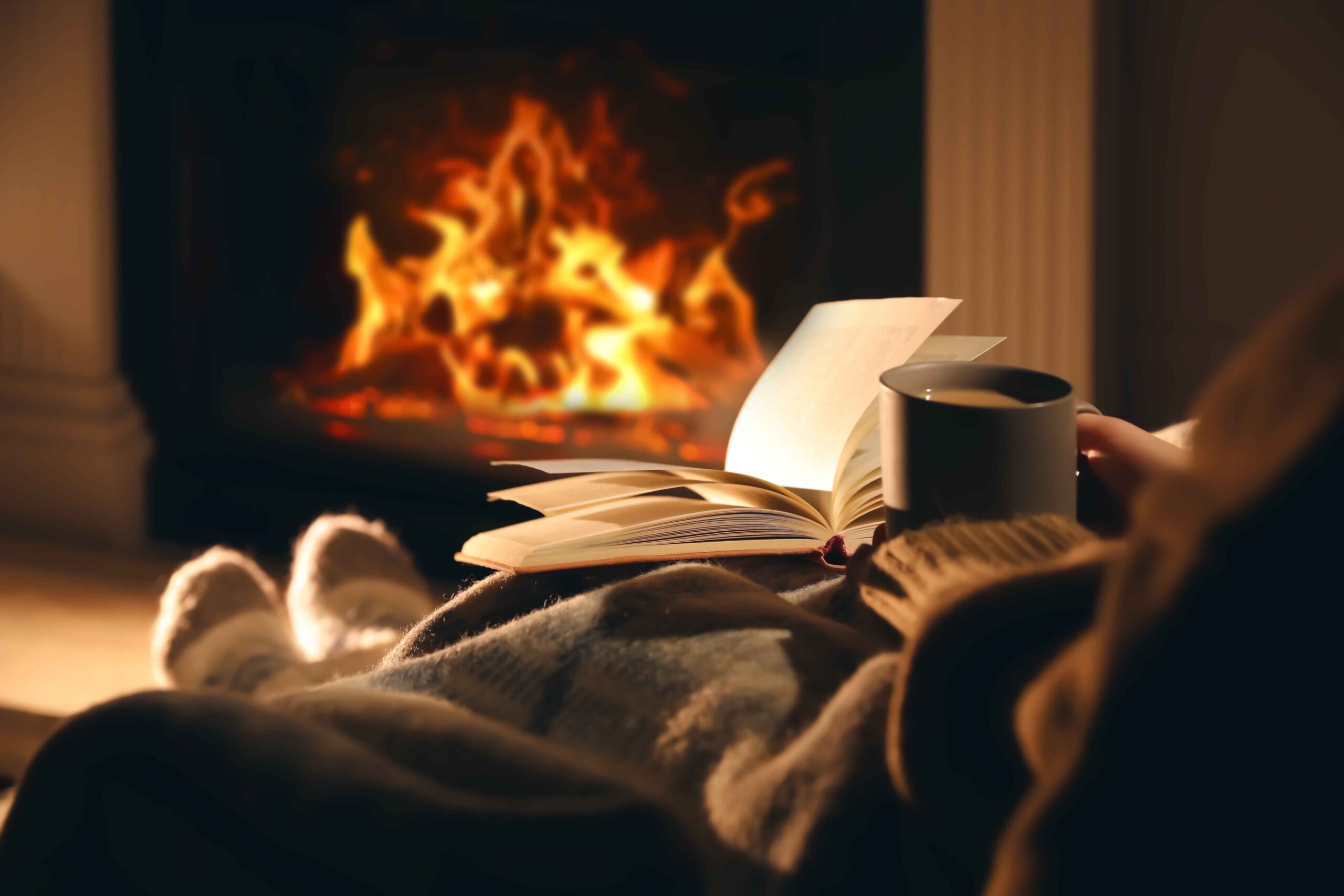 A person in a sweater sitting in front of a fireplace with a book and cup of coffee.